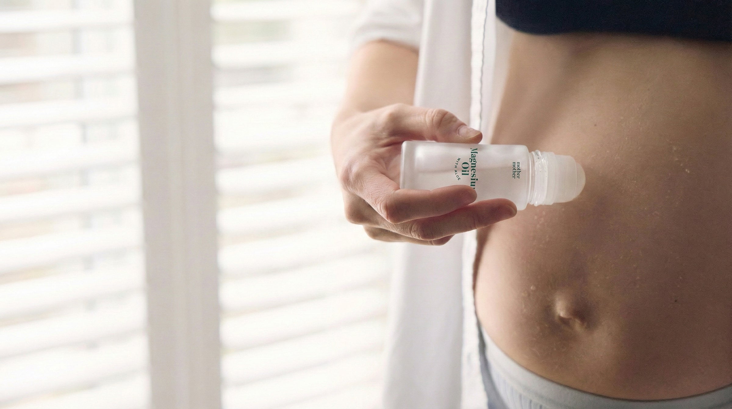 Person holding a bottle of magnesium oil near their stomach against a neutral background