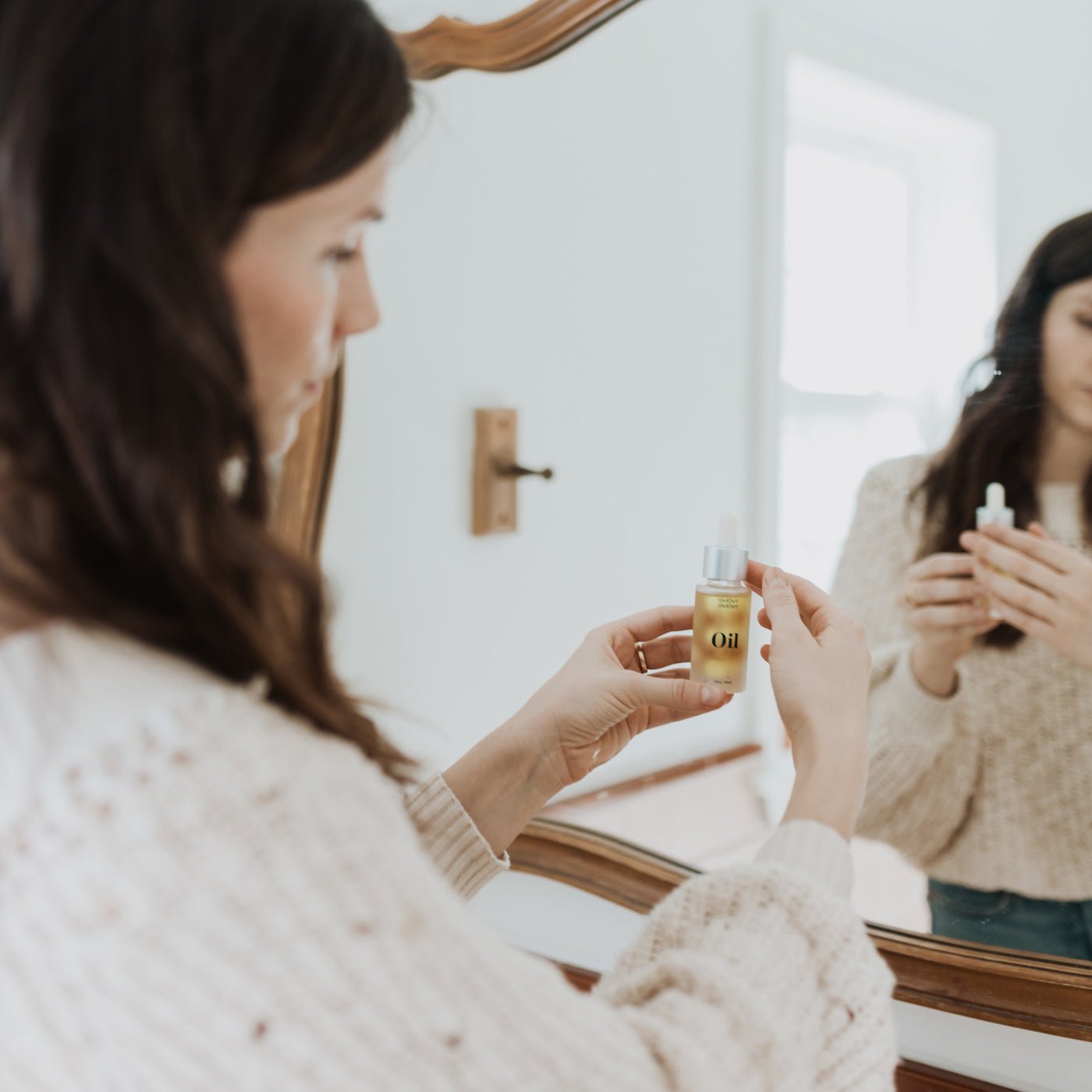woman with brown hair and beige sweater holding the oil bottle in front of her mirror